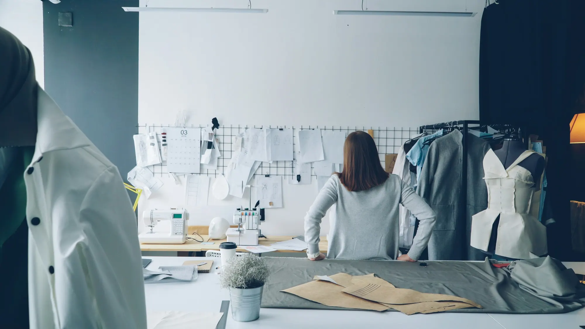 A designer standing at a cutting table over grey cloth and paper pattern pieces, with sketches pinned to the wall behind